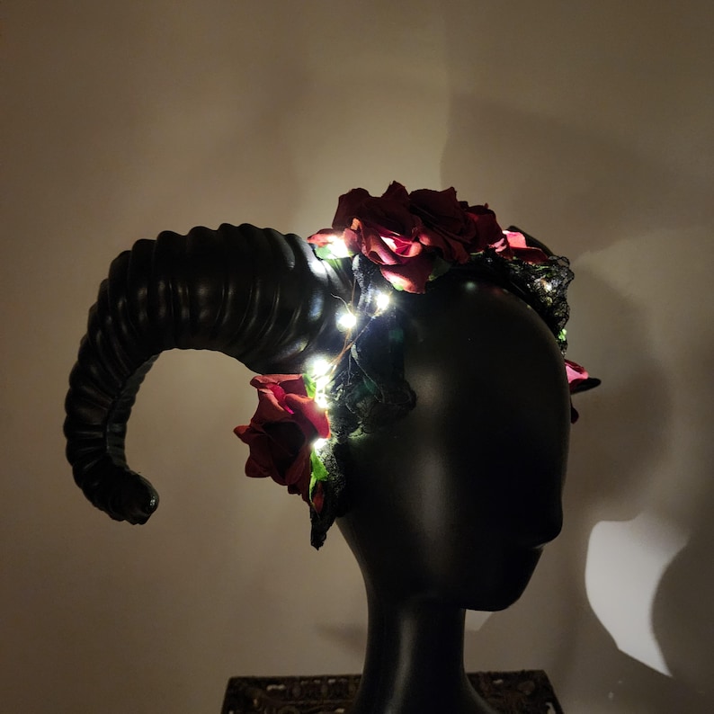 May include: A headpiece featuring large, curved, black ram horns and deep red artificial roses. The accessory is adorned with thin, glowing white LED fairy lights woven throughout the flowers and dark foliage, creating a luminous effect.