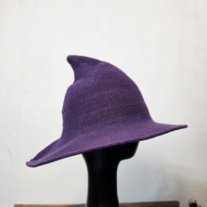 May include: A purple knitted witch hat with a pointed crown and a wide, slightly curved brim. The hat features a consistent, fine-knit texture throughout its surface, creating a structured yet flexible shape that tapers to a soft, rounded point.