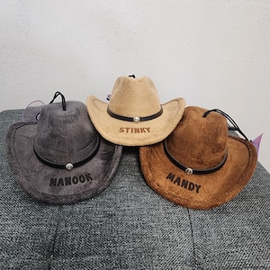 May include: Three cowboy hats in gray, tan, and brown. Each hat has a black band with a silver-colored jewel and a chin strap. The hats have the names "NANOOK", "STINKY", and "MANDY" embroidered on the front.