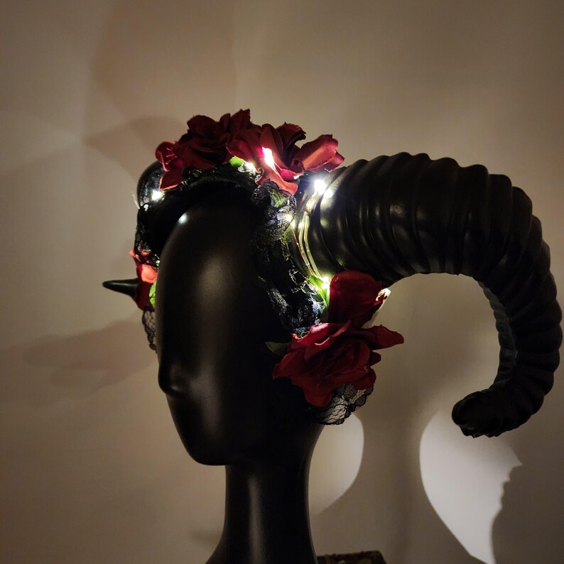 May include: A black headband featuring large, curved, ribbed ram horns, adorned with deep red artificial roses and black lace trim. Small, bright white LED lights are woven throughout the floral arrangement, providing illumination to the dark headpiece.