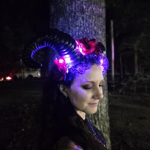 May include: A headpiece featuring large, curved black horns adorned with dark red fabric roses and black lace. The accessory is illuminated by small, bright purple LED lights integrated into the base of the horns and the floral arrangement.