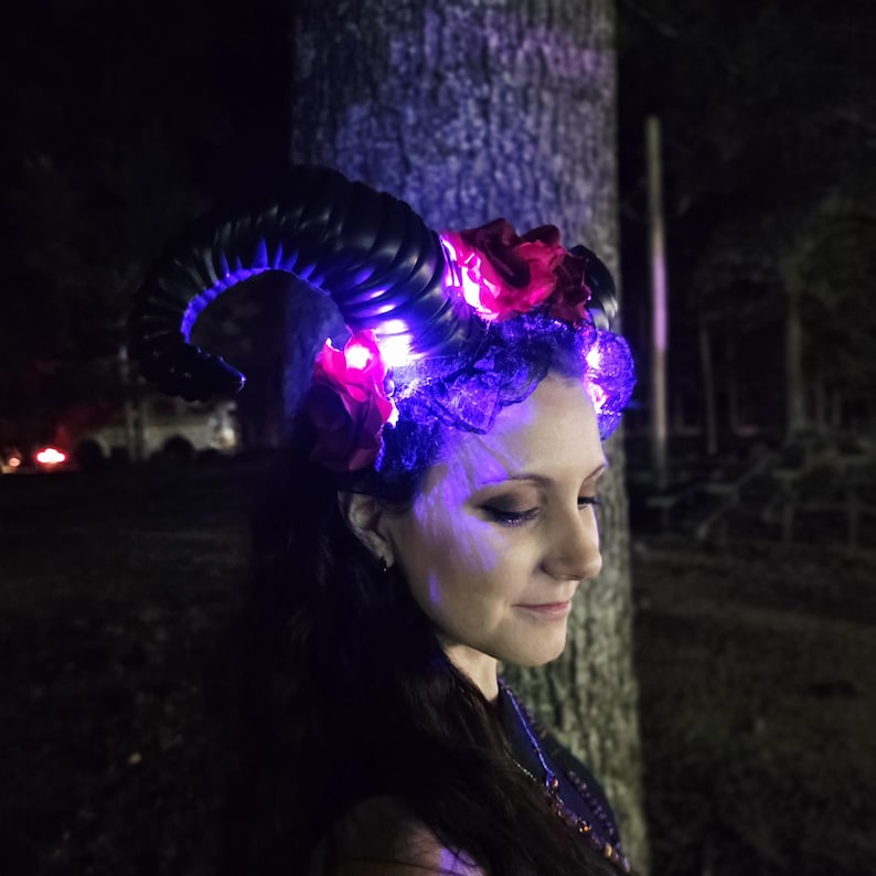 May include: A headpiece featuring large, curved black horns adorned with dark red fabric roses and black lace. The accessory is illuminated by small, bright purple LED lights integrated into the base of the horns and the floral arrangement.