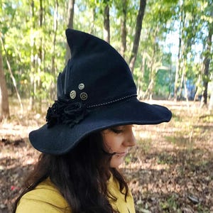 May include: A black witch hat with a pointed crown, wide brim, and decorative accents. The hat features black fabric roses, feathers, and small metallic gears. A decorative cord trims the brim.