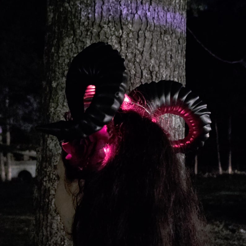 May include: A headband featuring two large, curved black ram horns with a textured, ridged surface. The base of the horns is illuminated by bright pink LED lights that cast a glow onto the dark, curled hair underneath the headpiece.