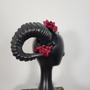 May include: A headpiece featuring a large, curved, black ram horn with a ribbed texture. The base of the horn is adorned with black lace fabric and two deep red artificial roses, one positioned at the top and one at the bottom of the lace.