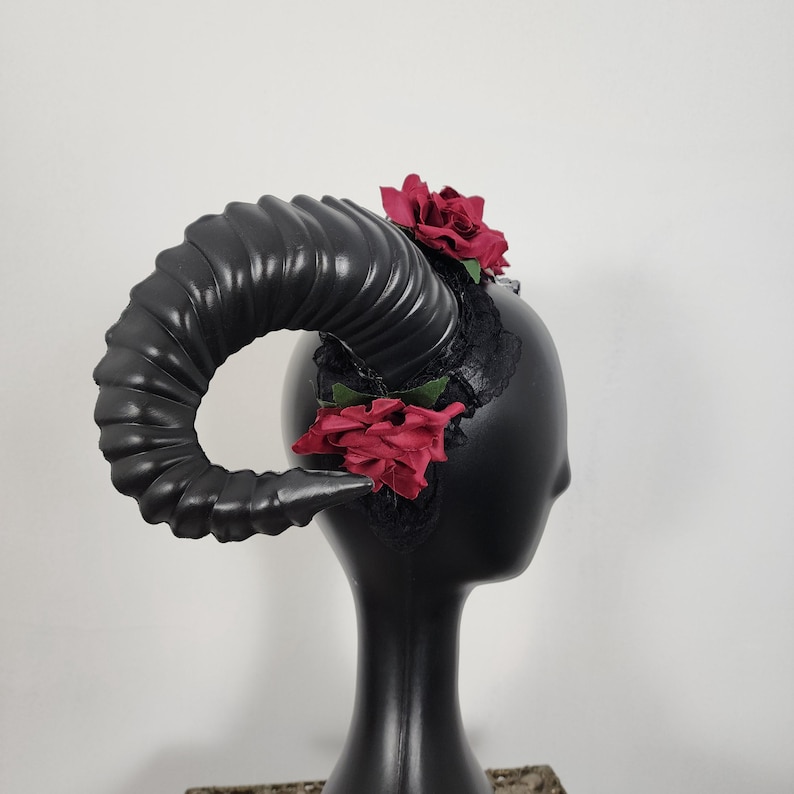 May include: A headpiece featuring a large, curved, black ram horn with a ribbed texture. The base of the horn is adorned with black lace fabric and two deep red artificial roses, one positioned at the top and one at the bottom of the lace.