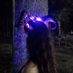 May include: A headpiece featuring two large, curved black ram horns adorned with red floral accents and integrated bright blue LED lights that illuminate the base of the horns and the surrounding area with a vibrant glow in the dark.