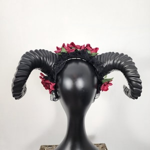 May include: A black headband featuring two large, curved, ridged ram horns extending from the sides. The headband is decorated with dark red fabric roses and black lace trim, positioned across the top and sides of the black mannequin head.