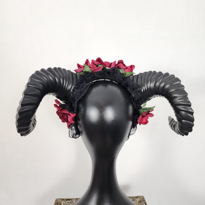 May include: A black headband featuring two large, curved, ridged ram horns extending from the sides. The headband is decorated with dark red fabric roses and black lace trim, positioned across the top and sides of the black mannequin head.