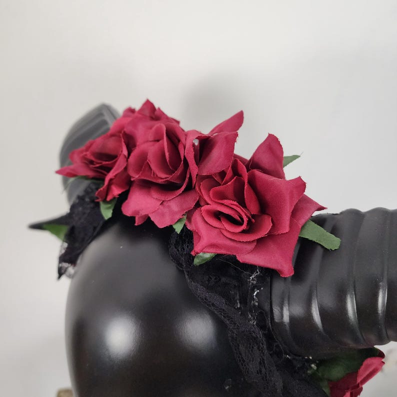 May include: A headband featuring large, deep red fabric roses and black lace trim. The accessory includes two prominent, curved black horns with a ribbed texture extending from the sides, creating a bold and structured headpiece design.