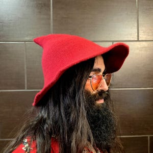 May include: A bright red knit hat with a pointed crown and a wide, slightly upturned brim. The hat is worn over long dark hair and heart-shaped sunglasses with orange-tinted lenses, creating a distinct profile against a dark tiled background.