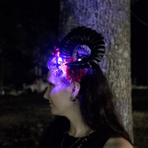 May include: A headpiece featuring a large, curved black ram horn and vibrant red artificial flowers. The accessory is illuminated by bright purple LED lights that cast a glow across the floral elements and the base of the horn structure.