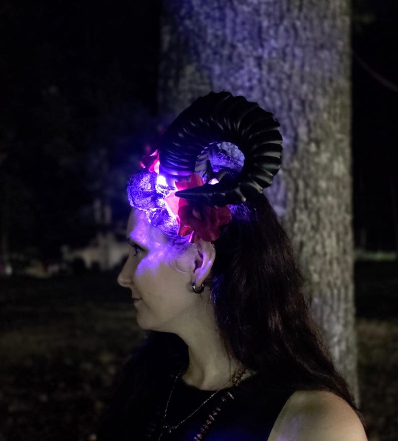 May include: A headpiece featuring a large, curved black ram horn and vibrant red artificial flowers. The accessory is illuminated by bright purple LED lights that cast a glow across the floral elements and the base of the horn structure.