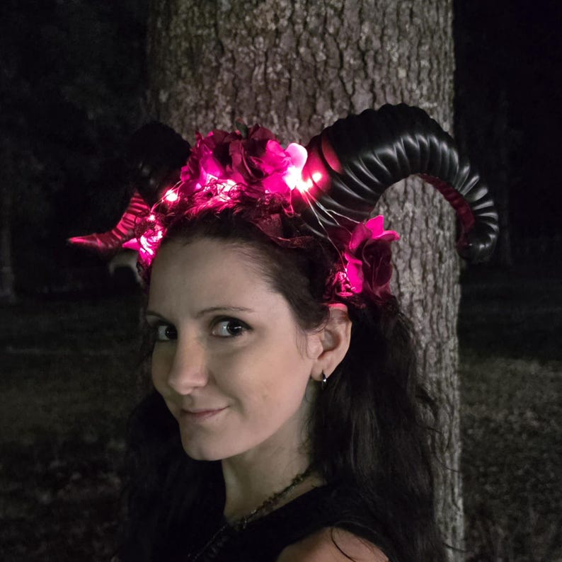 May include: A headband featuring two large, curved black ram horns and artificial dark red roses. The accessory is illuminated by small, bright pink LED lights woven throughout the floral arrangement, creating a glowing effect against the dark horns.