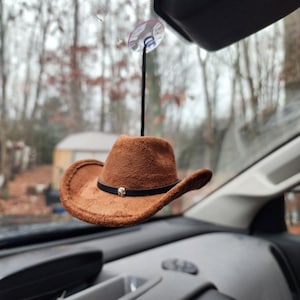 May include: A brown suede cowboy hat with a black band and a silver buckle hangs from a suction cup on a car's rearview mirror.
