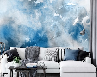 May include: A white sectional sofa with a gray throw blanket and blue and black patterned pillows in a living room with a blue and gray watercolor wall mural.