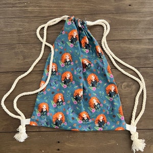 May include: A blue drawstring backpack featuring a repeating pattern of a cartoon character with red hair, surrounded by flowers and the word "BRAVE". The bag has white rope straps and tassels.