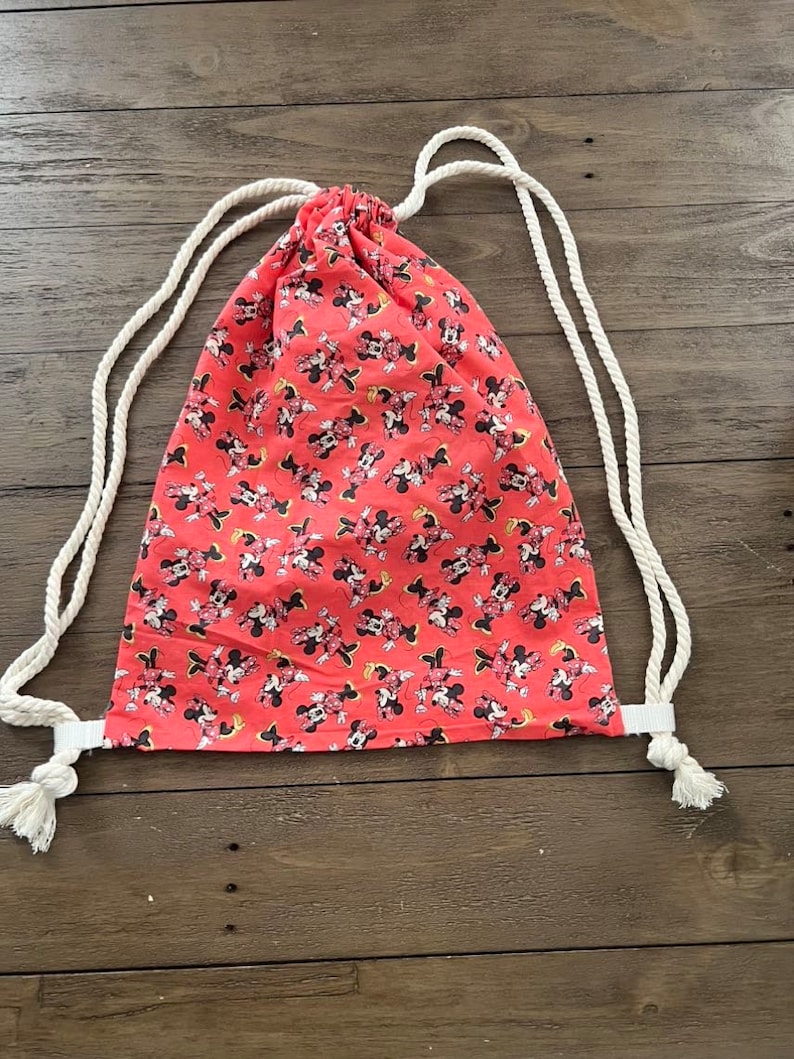 Puede incluir: Mochila con cord&oacute;n de color coral con un estampado repetido de Minnie Mouse. La bolsa tiene correas de cuerda blanca y una banda de tela blanca en la base de las correas. La bolsa est&aacute; hecha de tela ligera.