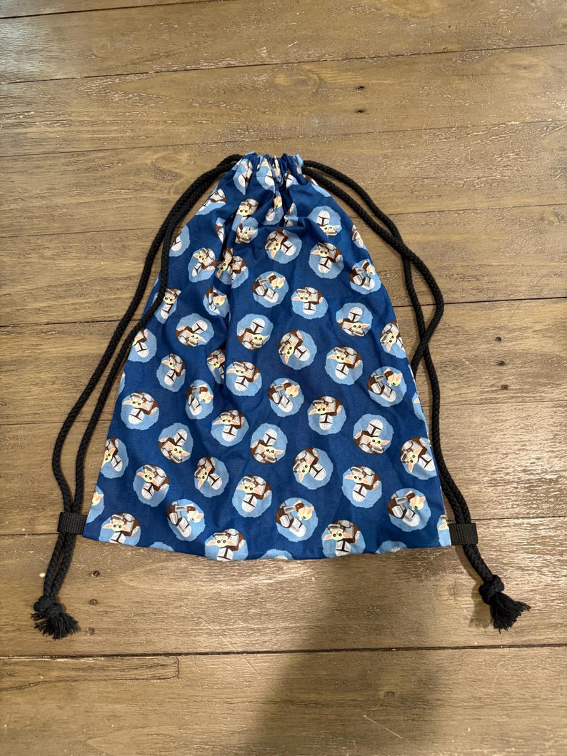 May include: A blue drawstring bag with a repeating pattern of brown teddy bears inside light blue circles. The bag has black drawstrings and is made of fabric.