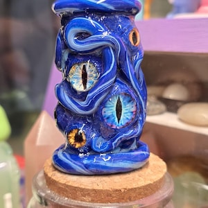 May include: A blue glass sculpture with multiple eyes, resembling an octopus or sea creature, sits atop a cork-topped jar. The sculpture has a swirling design with white accents and several eye-shaped details in shades of yellow and blue.