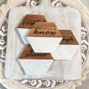 May include: Four hexagonal coasters with white marble tops and wooden bases. The coasters have inspirational quotes engraved on them: "Create with all your heart", "Be still and know", "Let your light shine", and "Peace is enough".