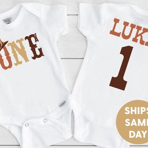 May include: Two white baby onesies with a cowboy theme. The front onesie has the word "ONE" in brown, orange, and yellow with a brown cowboy hat on top. The back onesie has the name "LUKA" in brown and the number "1" in brown.