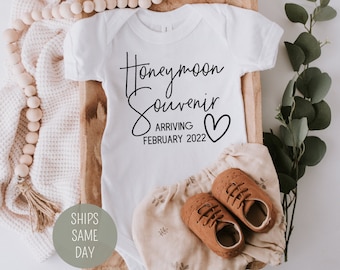 Puede incluir: Un body blanco para bebé con texto negro que dice "Souvenir de luna de miel Llegando febrero de 2022" con un pequeño corazón.