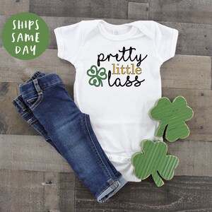 May include: White baby onesie with green shamrock and gold text that reads "pretty little lass". The onesie is laying on a wood surface with two green shamrock decorations.