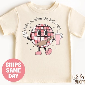 May include: A light beige t-shirt with a pink disco ball character holding a pink drink. The disco ball has a pink and white checkered pattern and the text "wake me when the ball drops" is written around it.
