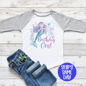 May include: A white and gray raglan shirt with a mermaid graphic and the text "Birthday Girl". The mermaid has blue hair and a teal tail. Blue jeans are folded next to the shirt.