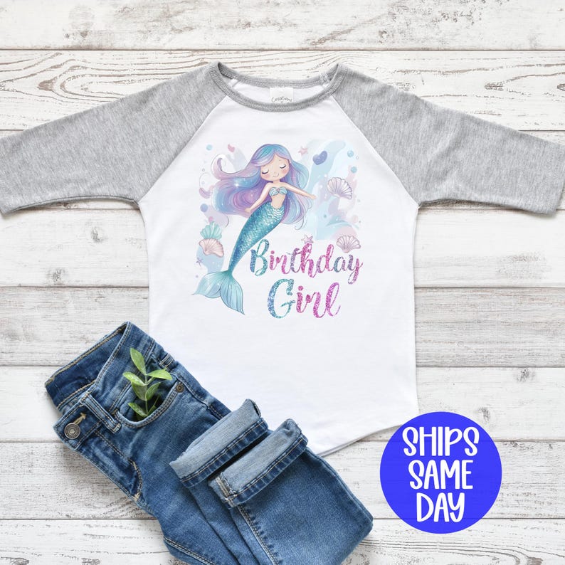 May include: A white and gray raglan shirt with a mermaid graphic and the text "Birthday Girl". The mermaid has blue hair and a teal tail. Blue jeans are folded next to the shirt.
