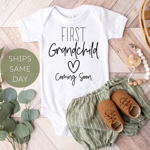 May include: White baby onesie with black text that reads "First Grandchild Coming Soon" with a heart symbol. The onesie is laying on a wooden surface with a pair of brown baby shoes and a green and white striped baby outfit.
