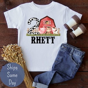 May include: White t-shirt with a red barn, a pig, a horse, and a cow. The number 2 with a black and white cow print is on the left side of the barn. The name "Rhett" is printed below the barn.