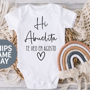 May include: White baby onesie with black text that reads "Hi Abuelita Te Veo En Agosto" with a heart symbol.