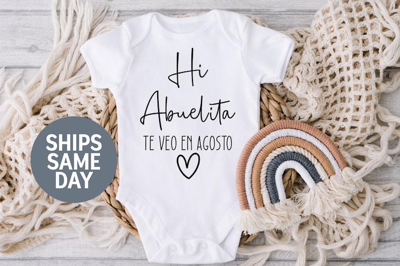 May include: White baby onesie with black text that reads "Hi Abuelita Te Veo En Agosto" with a heart symbol.