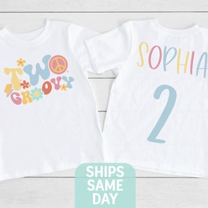 May include: Two white t-shirts with colorful graphics. One shirt says "Two Groovy" with a peace sign and flowers. The other shirt has the name "Sophia" and the number "2" in pastel colors. The shirts are likely for a second birthday.