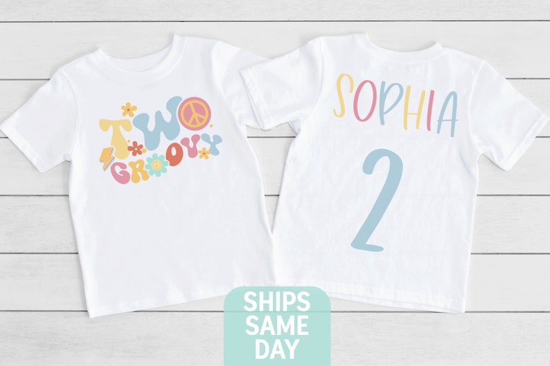 May include: Two white t-shirts with colorful graphics. One shirt says "Two Groovy" with a peace sign and flowers. The other shirt has the name "Sophia" and the number "2" in pastel colors. The shirts are likely for a second birthday.