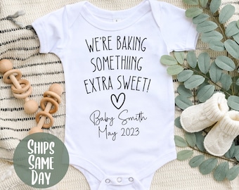 Può includere: Un body bianco per neonato con il testo "We're baking something extra sweet!" e un cuore. Sotto il testo si legge "Baby Smith May 2023".