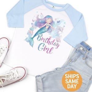 May include: White and light blue raglan shirt with a mermaid graphic and the text "Birthday Girl". The shirt is paired with light wash denim jeans and white sneakers.