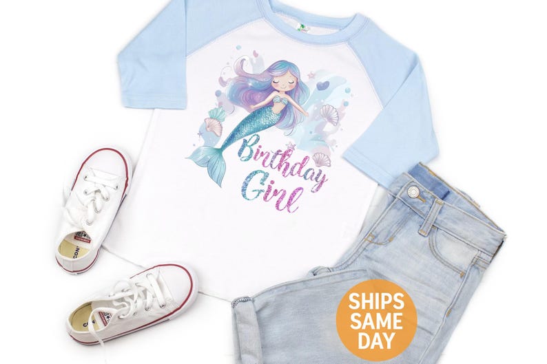May include: White and light blue raglan shirt with a mermaid graphic and the text "Birthday Girl". The shirt is paired with light wash denim jeans and white sneakers.