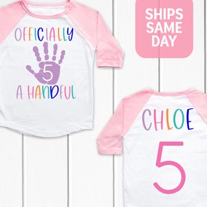 Pu&ograve; includere: Due magliette in stile baseball rosa e bianche. Una maglietta ha un'impronta di mano viola con il numero 5 all'interno e il testo "Ufficialmente una manciata". L'altra maglietta ha il nome "Chloe" in colori arcobaleno e il numero 5 in rosa.