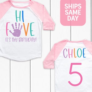 K&ouml;nnte beinhalten: Ein wei&szlig;es und pinkfarbenes Baseball-Shirt mit dem Text "Hi Five It's My Birthday!" in Regenbogenfarben und einem lila Handabdruck. Die R&uuml;ckseite des Shirts tr&auml;gt den Namen "Chloe" in Regenbogenfarben und die Zahl "5" in Pink.