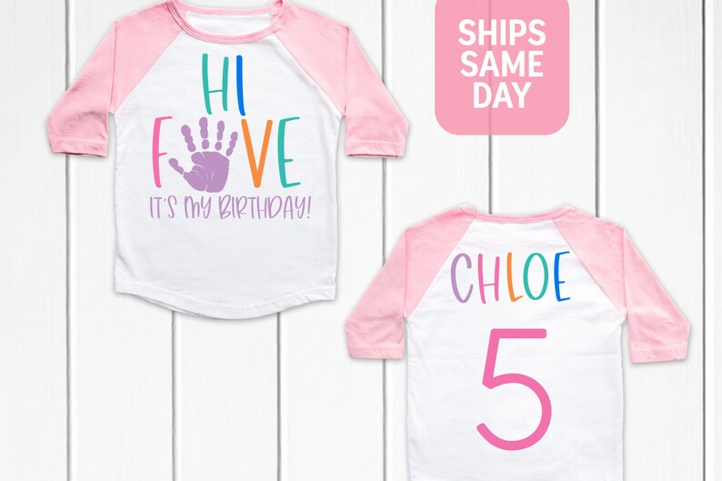K&ouml;nnte beinhalten: Ein wei&szlig;es und pinkfarbenes Baseball-Shirt mit dem Text "Hi Five It's My Birthday!" in Regenbogenfarben und einem lila Handabdruck. Die R&uuml;ckseite des Shirts tr&auml;gt den Namen "Chloe" in Regenbogenfarben und die Zahl "5" in Pink.