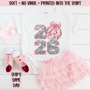 May include: White t-shirt with the numbers "2026" in silver glitter and a disco ball graphic. The shirt is paired with a pink tutu skirt and pink shoes. The text "SOFT • NO VINYL • PRINTED INTO THE SHIRT" is printed at the top.