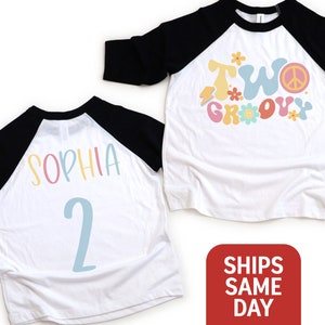 May include: White and black raglan style shirt with the words "TWO GROOVY" in pastel colors and a peace sign. The back of the shirt has the name "SOPHIA" and the number "2" in pastel colors. A fun birthday shirt.