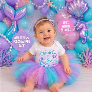 May include: A white short-sleeved bodysuit with a graphic print featuring a mermaid tail, a seashell, and the text One-der the Sea, paired with a layered tulle tutu in shades of purple and blue, and a matching headband with a small starfish.
