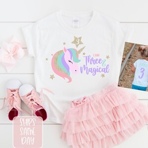May include: White t-shirt with a unicorn design and the text "I am Three & Magical". The back of the shirt has the number "3" in purple glitter. The shirt is paired with a pink tulle skirt and pink sneakers.