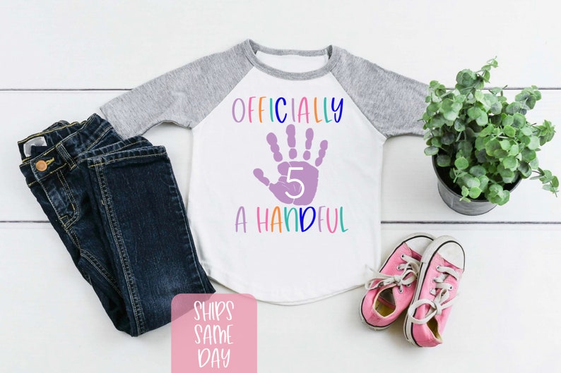Pu&ograve; includere: Una maglietta a maniche lunghe bianca e grigia con il testo "Ufficialmente una manciata" e un'impronta di mano viola con il numero 5 all'interno.