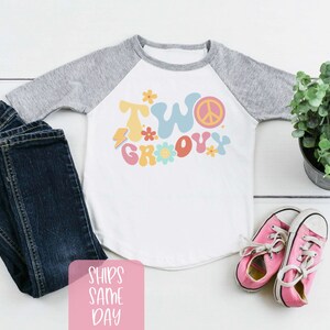 May include: A white and gray raglan style toddler shirt with the words "TWO GROOVY" in colorful retro font, paired with dark blue jeans and pink sneakers. A small potted plant is also in the image.