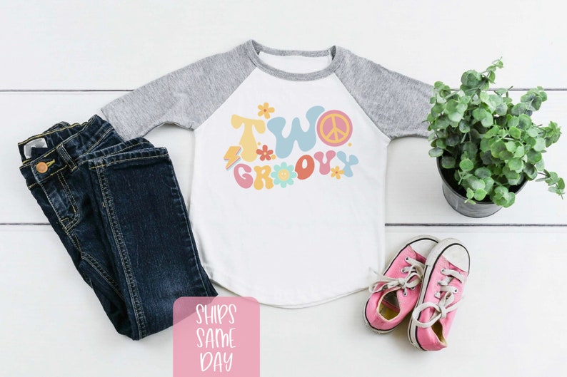 May include: A white and gray raglan style toddler shirt with the words "TWO GROOVY" in colorful retro font, paired with dark blue jeans and pink sneakers. A small potted plant is also in the image.